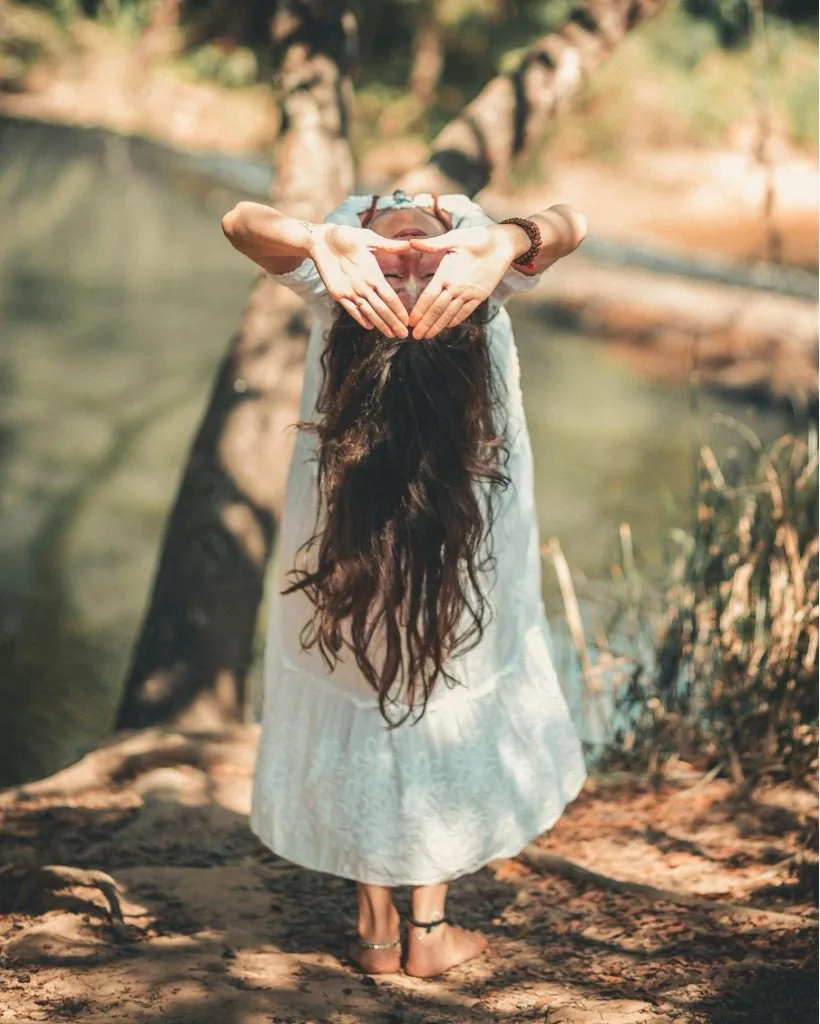 Liis Ugandi in a forward fold outdoors by the water, hands over her face, white dress. Goyoga Tallinn.