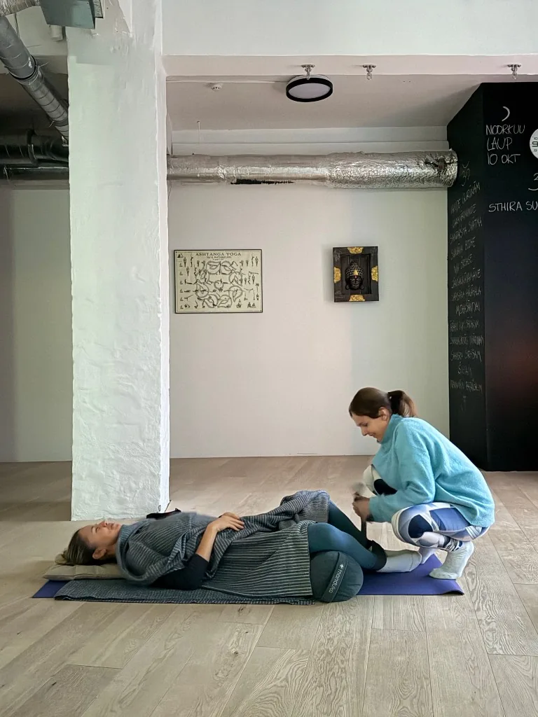 Liis Ugandi in a light blue fleece, crouching to adjust a student in a supported restorative pose at Goyoga Tallinn.