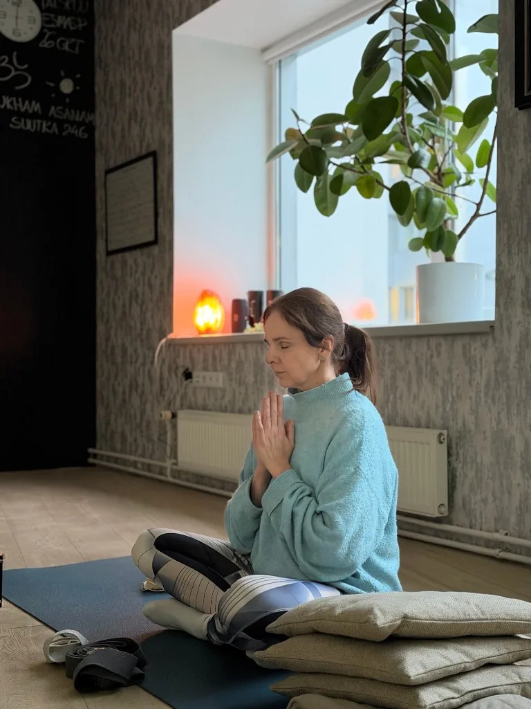 Liis Ugandi seated with hands together, light blue sweater, bolsters and a salt lamp at Goyoga Tallinn.